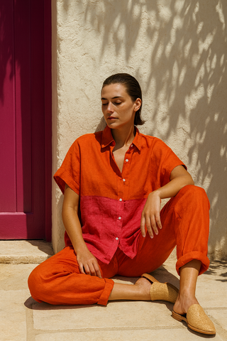 Woman sitting on a stone floor wearing a two-tone orange and pink outfit with a pink wall and shadow in the background.