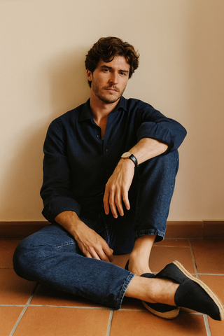 Man sitting on a tiled floor wearing a navy shirt and jeans against a beige wall.