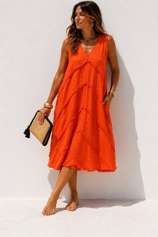 A woman wearing a sleeveless bright orange midi dress with a V-neckline and tiered ruffle details, holding a woven clutch bag against a white wall.
