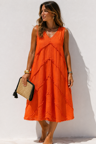 A woman wearing a sleeveless bright orange midi dress with a V-neck and tiered ruffle details, holding a woven clutch bag against a plain white wall.