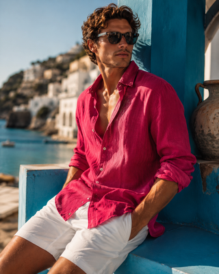 A man wearing a bright pink linen button-down shirt and white shorts sits on a blue wall by the sea, wearing dark sunglasses.